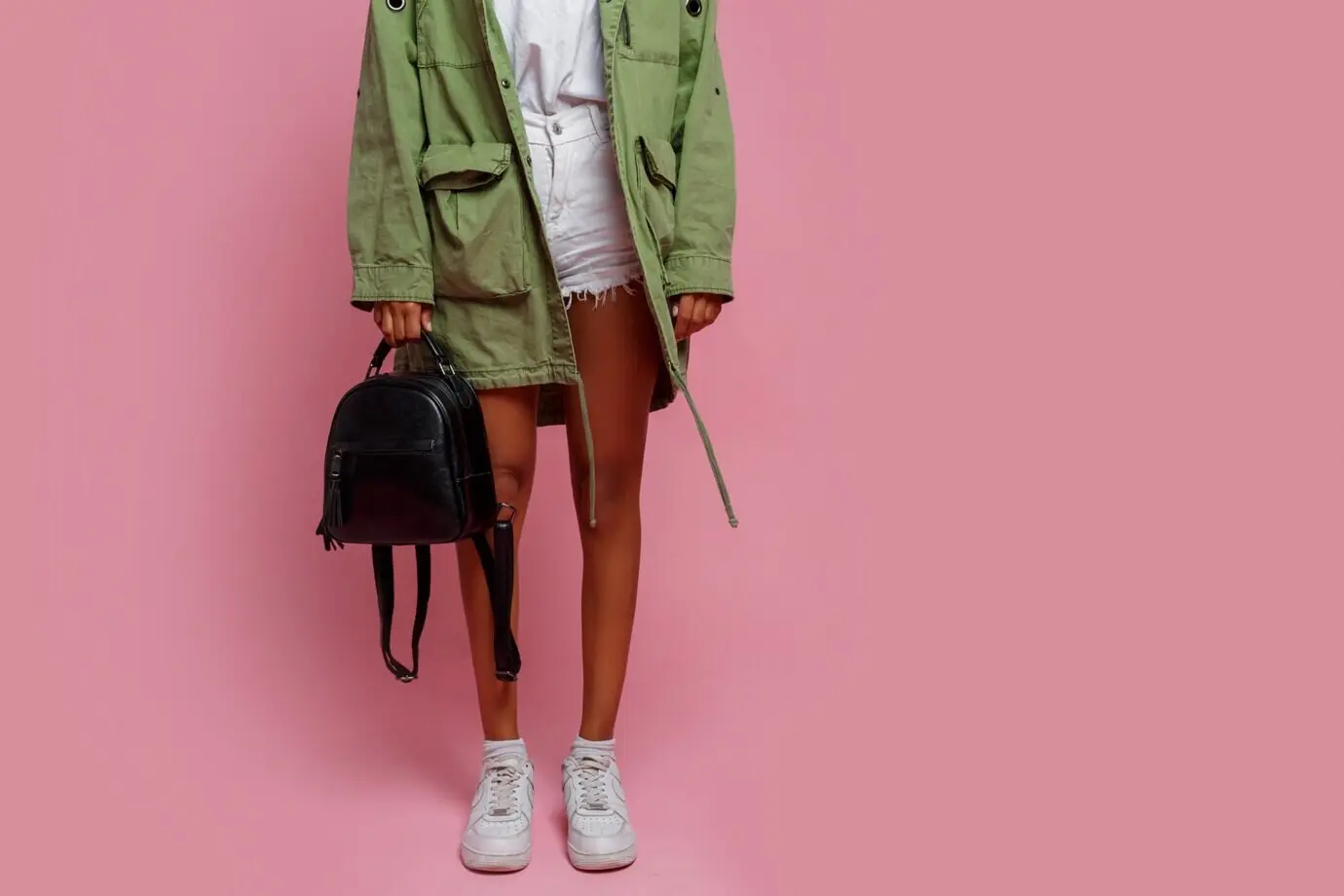 Modedetails. Schwarze Frau in grüner Jacke, weißen Shorts und Sneakers steht vor pinkem Hintergrund im Studio.